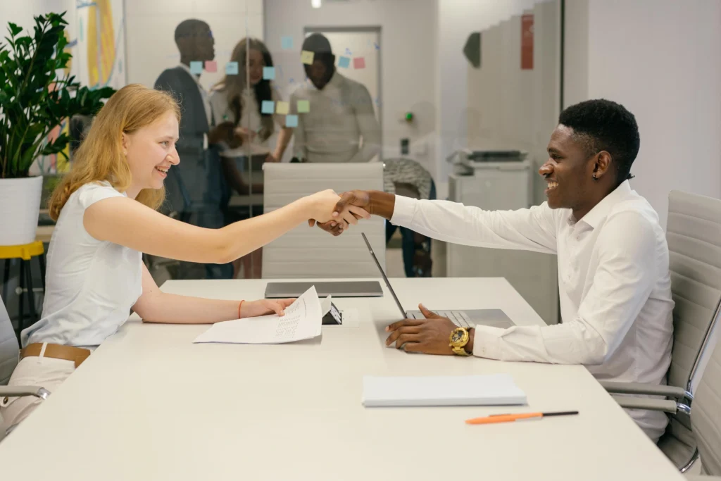 Two colleagues shaking hands across a desk with a team collaborating in the background